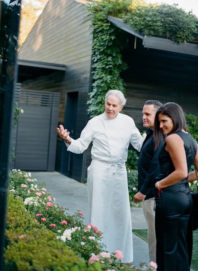 A chef in whites speaking with guests beside the rose beds